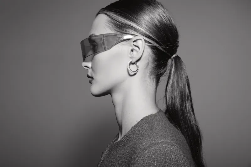 Profile of a white woman with pale skin wearing futuristic sunglasses and hoop earrings against a plain grey background.
