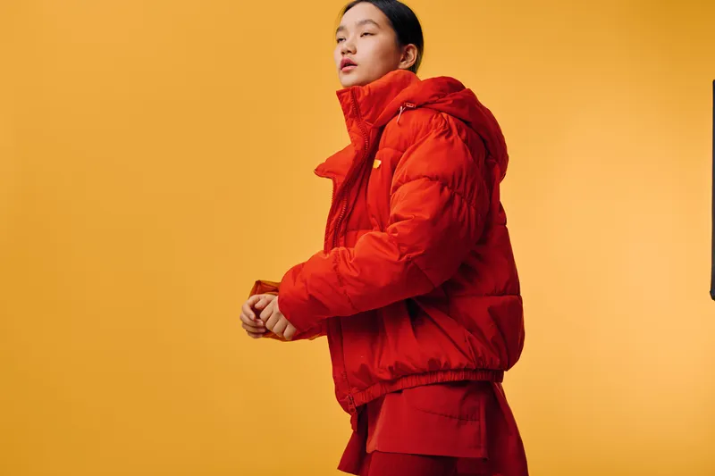 Woman wearing red puffer jacket and red outfit standing against orange background. Fashionable female model in warm winter clothing, casual style with confident expression in studio.