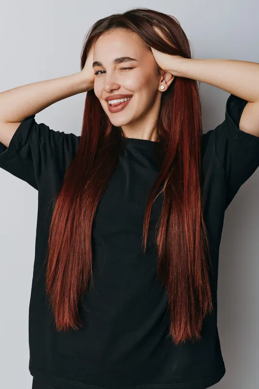 Woman with long red hair wearing black sweater posing with hands behind head and winking on gray background. Portrait of happy female smiling and showing playful expression indoors.