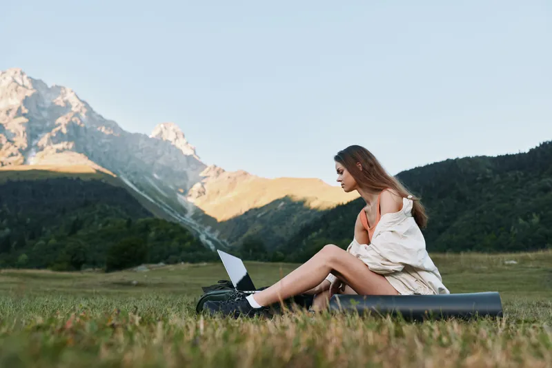 A focused woman sits on a grassy field with a laptop, enjoying a sunny day outdoors. She works remotely, surrounded by mountains, feeling productive and inspired.