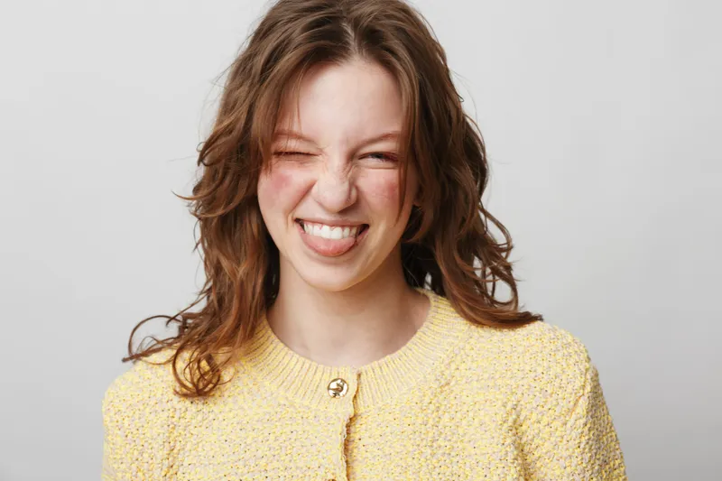 Young woman with curly brown hair wearing yellow sweater makes playful face sticking out tongue. Studio portrait on light background. Concept of fun, youth, and carefree attitude.