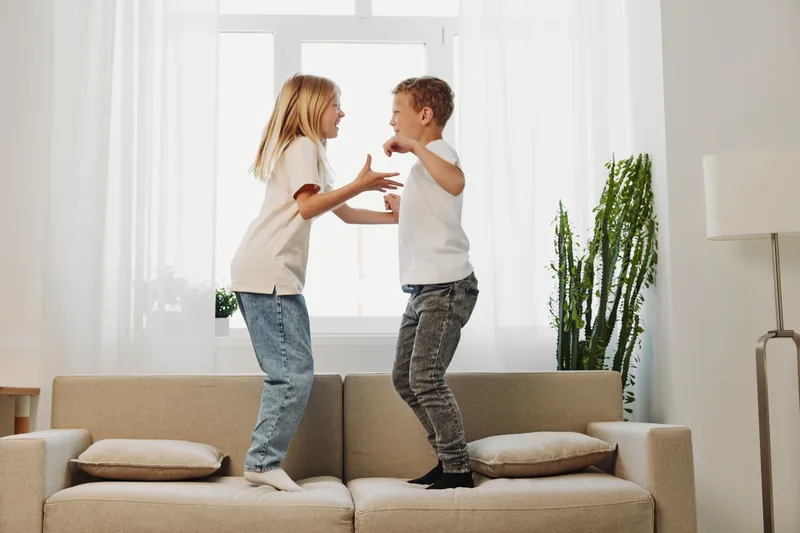 Two happy children jumping on sofa in bright living room, wearing casual clothes and enjoying playful moment. Concept of childhood fun and family bonding.
