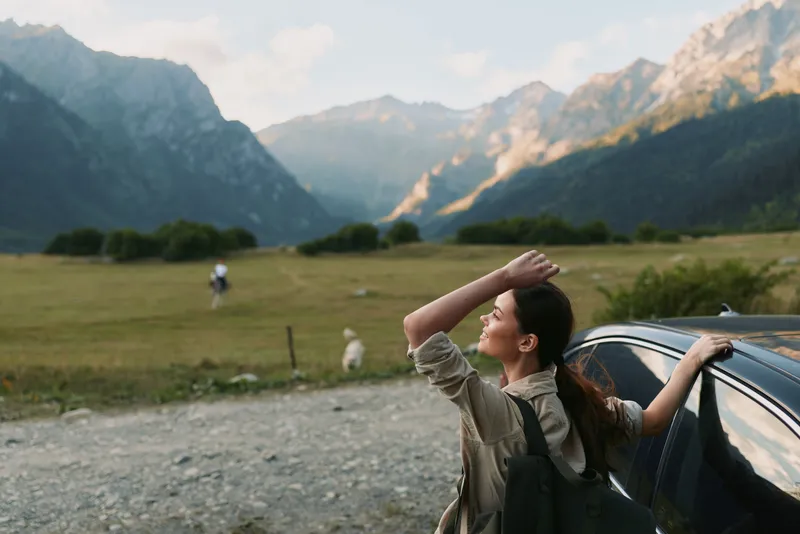 Woman beside a car in a mountain meadow, enjoying fresh air and wide landscape, conveys travel freedom and outdoor lifestyle in a sunny rural scene