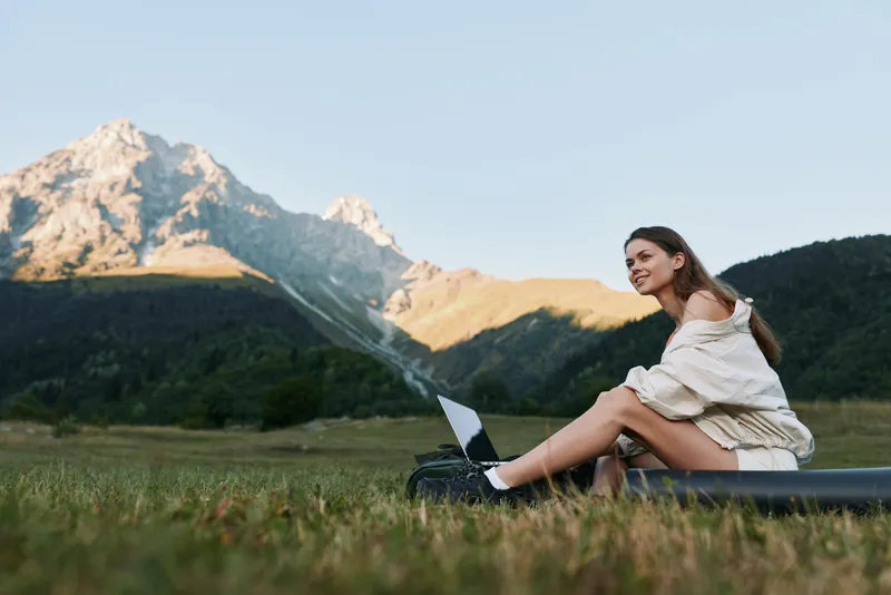 A cheerful woman sits on a grassy hill with a laptop, enjoying a sunny day in a mountain landscape. She smiles, focused on her work, creating a calm outdoors office atmosphere.