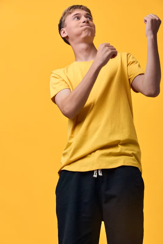 Energetic young male in a yellow shirt raises fists in a celebratory pose against a vivid orange background, bold studio shot emphasizing motion, mood, and athletic confidence