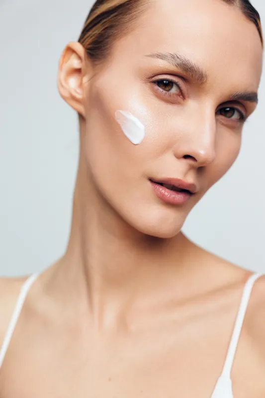 A close-up portrait of a young woman with clear skin applying white cream on her cheek. The image conveys skincare, beauty routine, hydration, and natural complexion concepts.