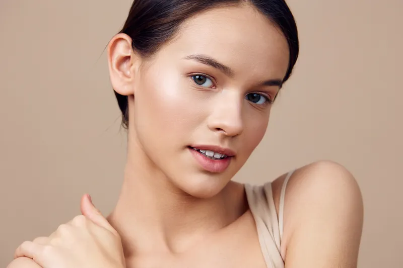 Natural beauty portrait of a young woman with flawless skin, wearing a light top against a neutral background, conveying confidence and tranquility