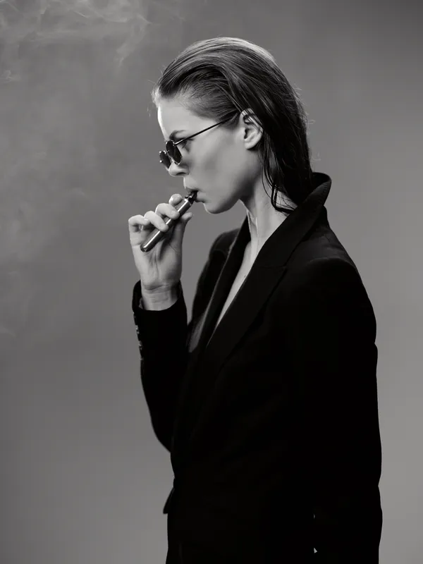 Black and white portrait of a woman vaping in dark stylish clothing with sunglasses, exhaling smoke in a minimalistic studio setting.