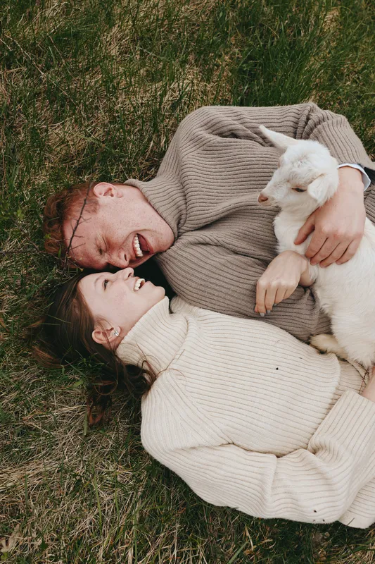 Joyful couple outdoors, lying on grass with a curious goat kid, warm sweaters, candid affection, sunlit moment, natural colors, relaxed pose, countryside atmosphere