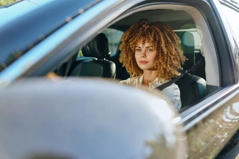 Young woman driving a car with curly hair and wearing a seatbelt, sitting in a modern vehicle interior on a sunny day, focused and calm expression.