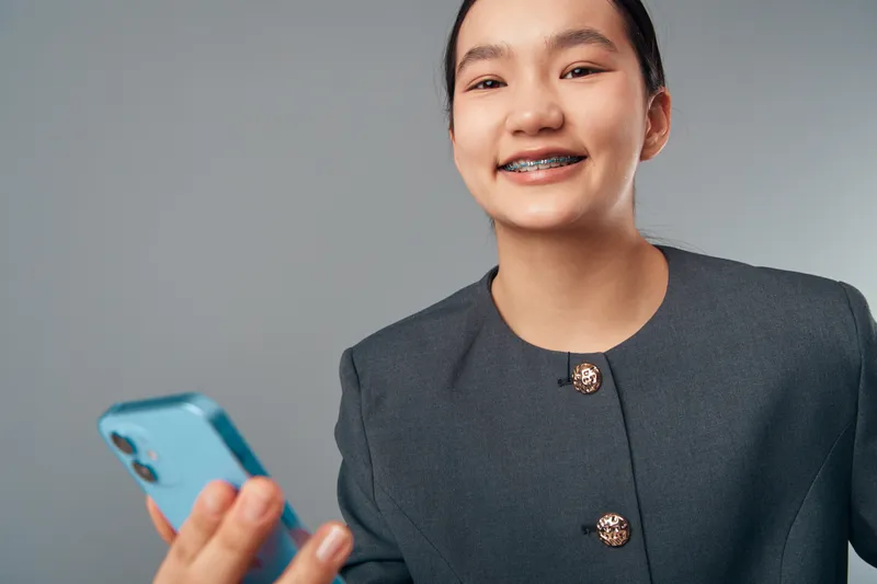 Teenage girl with braces holding smartphone smiling to camera wearing formal jacket on gray background. Young female student with modern device and casual hairstyle looking confident and happy.