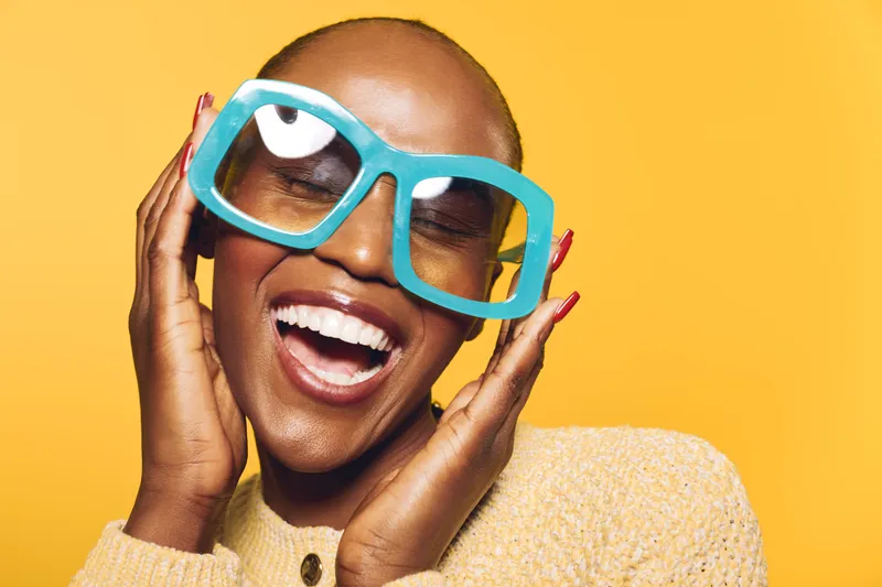 Happy African American woman with dark skin wearing large blue glasses and a beige sweater smiles with closed eyes on yellow background. Concept of joy, fashion, and beauty.