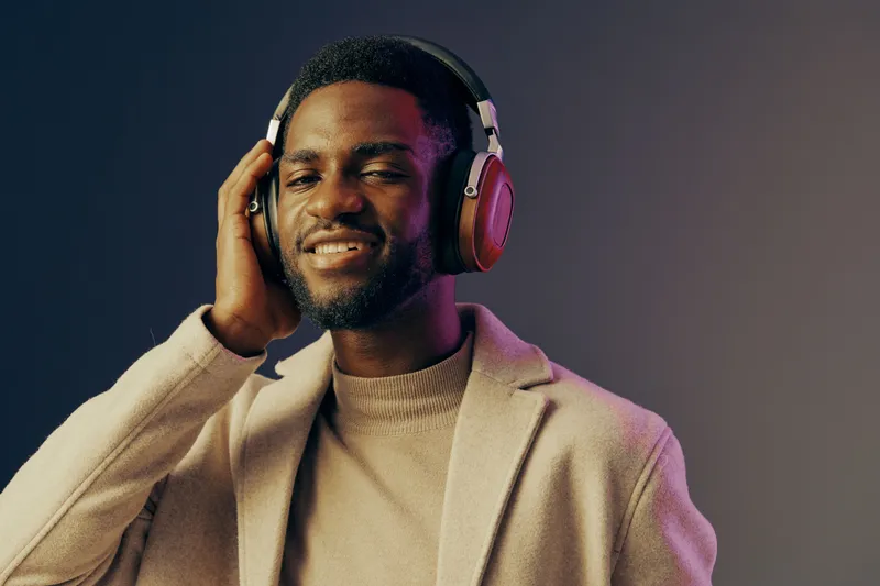 Young Black man enjoying music with headphones, dressed in a stylish beige coat against a gradient background, conveying joy and relaxation