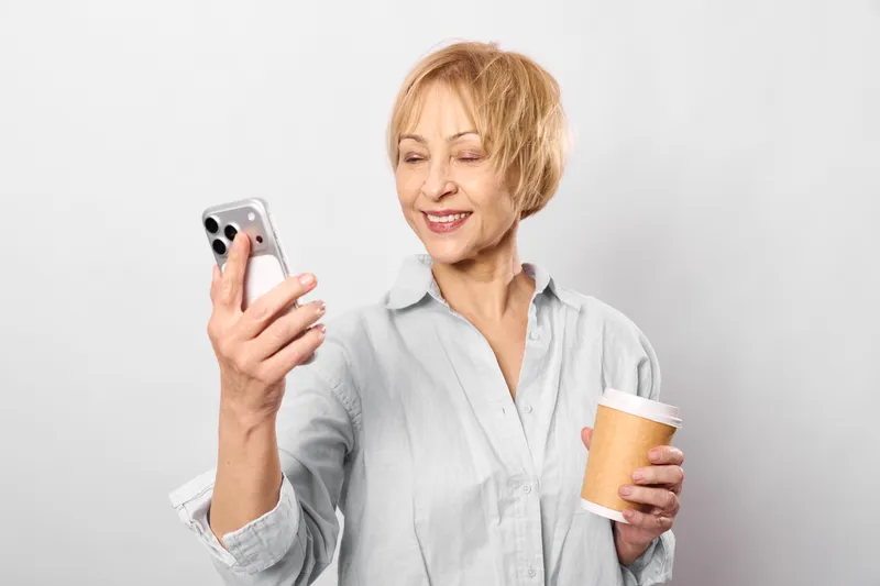 Senior woman holds a smartphone and a coffee cup, eyes closed with a tense expression, showing stress and fatigue in daily life, concept for burnout and mental health.