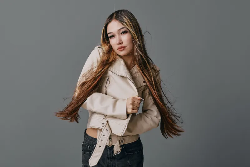 Young woman with long brown hair wearing beige jacket and black jeans posing against gray background. Casual style, confident expression, modern fashion and studio portrait.