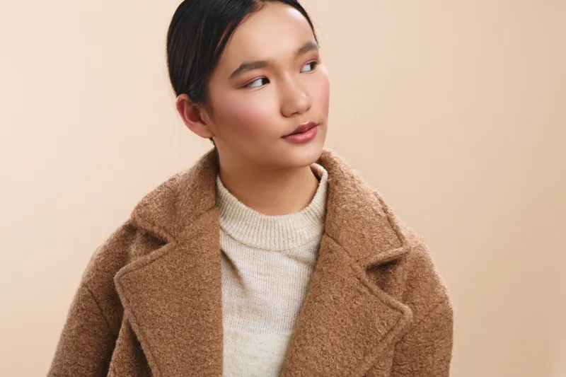 Asian woman wearing a warm tan coat stands against a neutral background, presenting a calm and stylish mood. She looks away with a thoughtful expression, conveying poised confidence and modern