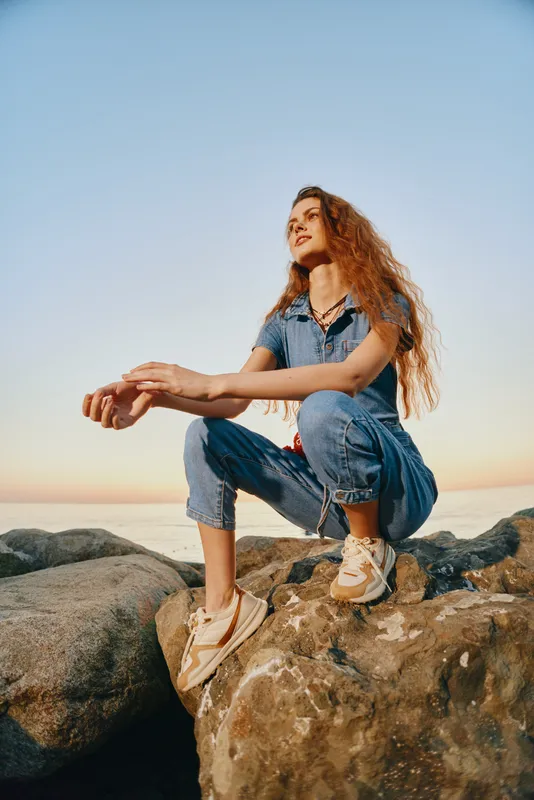 Lifestyle woman in modern boho-western denim street style posing on rocks with film color tones during sunset, embodying relaxed and confident mood outdoors.