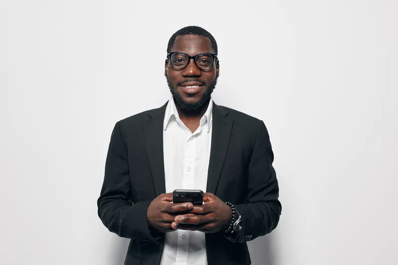 African American man with dark skin wearing glasses and black suit smiling while holding smartphone in hands against white background in studio portrait.