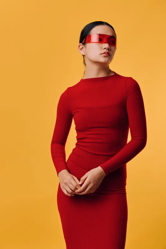 Young woman in red fitted dress and futuristic red visor stands against orange background, posing with confidence and modern fashion style in high key studio setting.