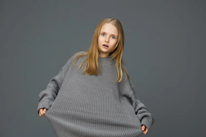 A surprised young girl with long hair in an oversized gray sweater, set against a muted background, expressing curiosity and innocence in a cozy atmosphere