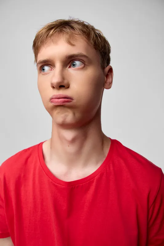 Man in a red shirt displays a puzzled expression, eyes turned aside, conveying confusion and curiosity in a casual studio setting, suitable for lifestyle or fashion use