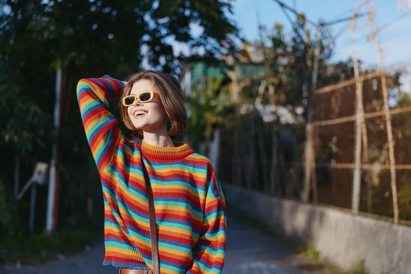 Woman in a colorful striped sweater and sunglasses smiles broadly on a sunny street, casual fashion portrait with short hair, joyful expression and relaxed urban vibe.