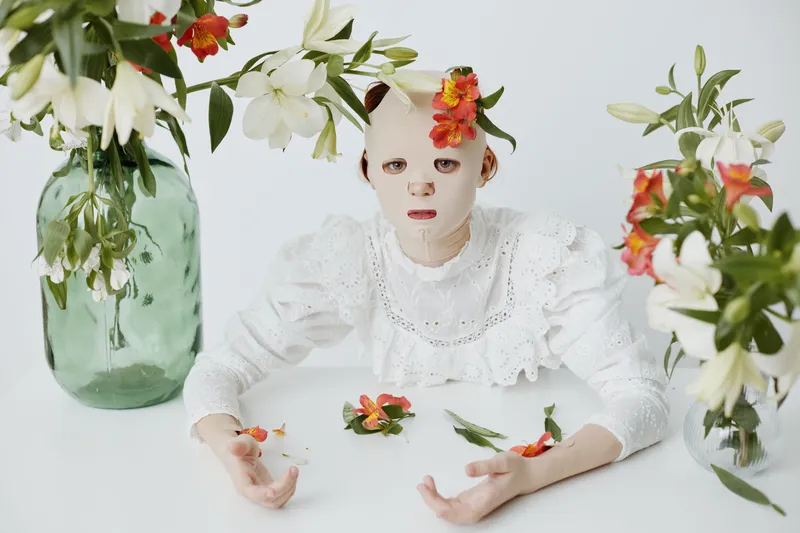 Artistic conceptual portrait of a masked model surrounded by flowers, exploring themes of identity and beauty amidst a serene, bright atmosphere