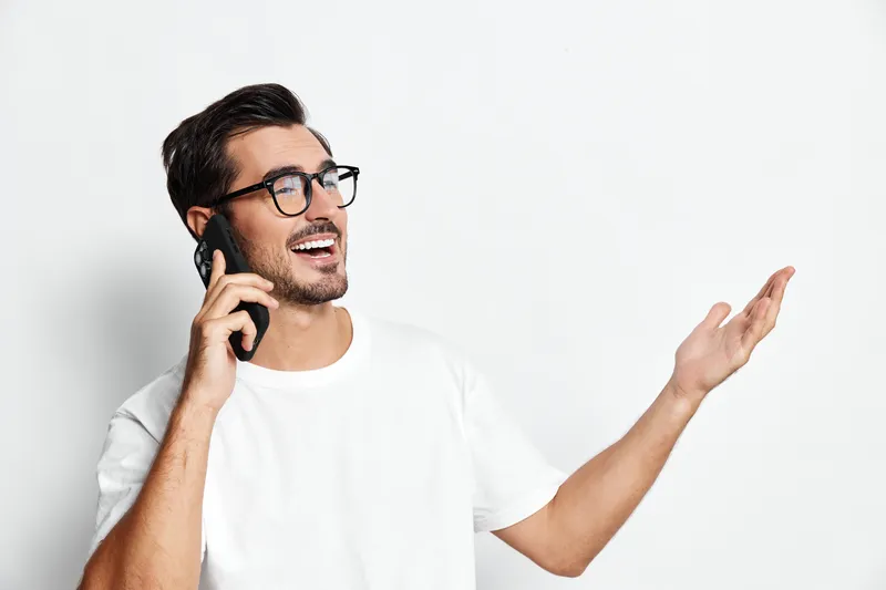Smiling cheerful young caucasian man talking on mobile phone, expressing joy while gesturing with his hand, isolated on a minimalistic light background.