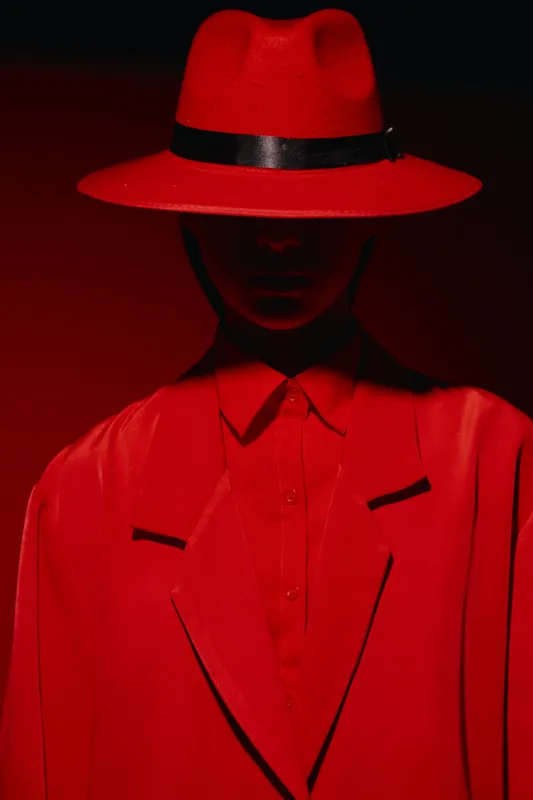 Mysterious fashion model wearing an oversized red suit and wide-brimmed hat with a shadowed face. Creative beauty shot with bold color and dramatic lighting.