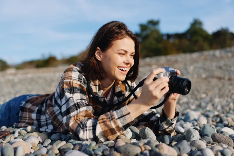 Woman photographer lying on a pebble beach, smiling while shooting with a camera. Casual plaid jacket, clear sky, vivid natural light enhances outdoor photography.