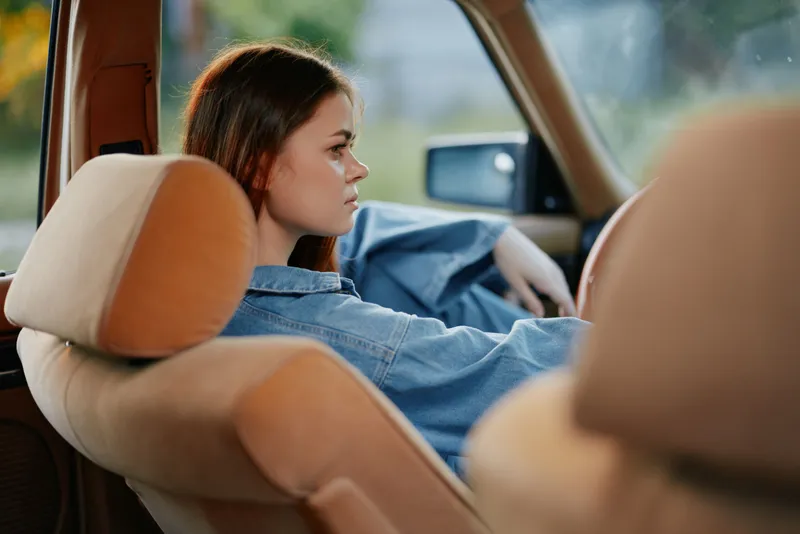 Young woman in denim shirt relaxing in a car interior, conveying a calm and contemplative mood with warm colors in the background.