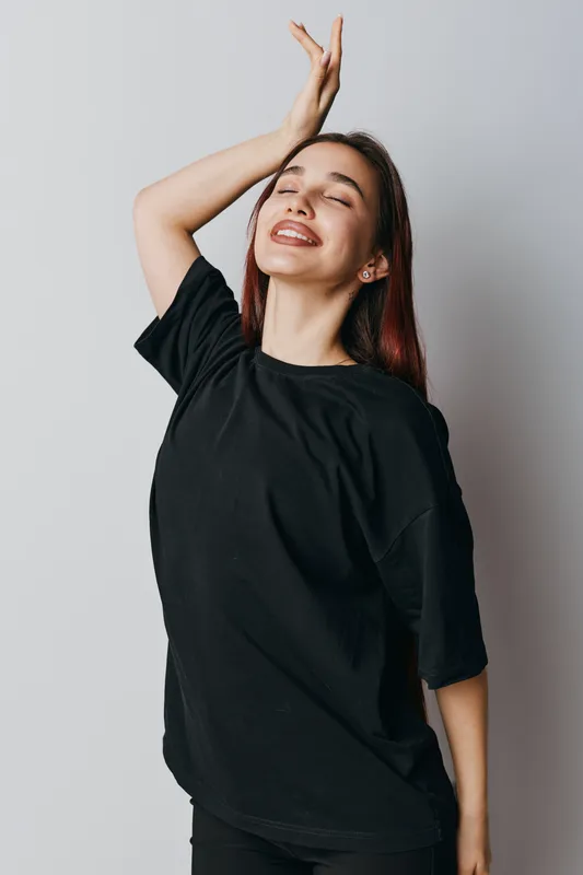 Young woman in black t shirt with closed eyes smiling and hand raised posing against plain background. Female model with long hair enjoying moment in casual clothing and relaxed mood.