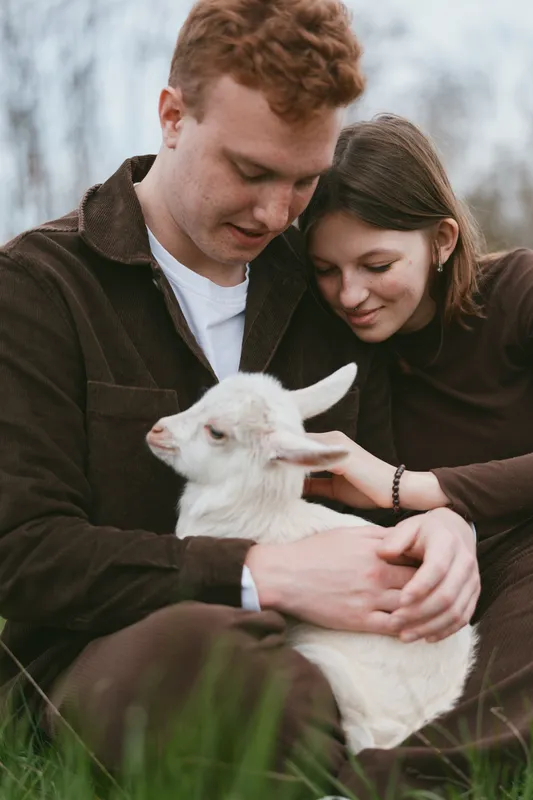 Young couple gently holds a white baby goat while sitting outdoors in casual clothes, sharing a tender moment of connection and care in a natural lifestyle setting.