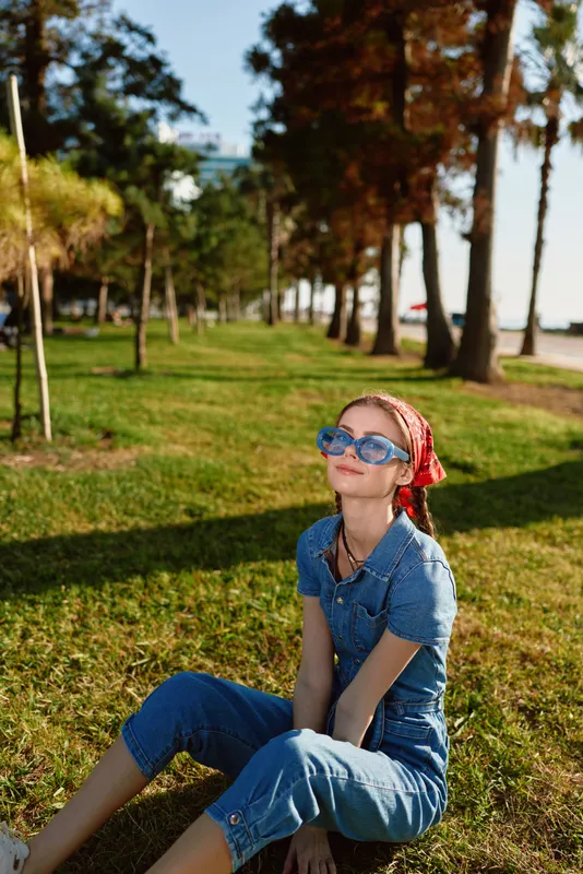 Lifestyle woman in modern boho-western denim street style wearing sunglasses, sitting on grass with film color effect and relaxed expression under autumn light.