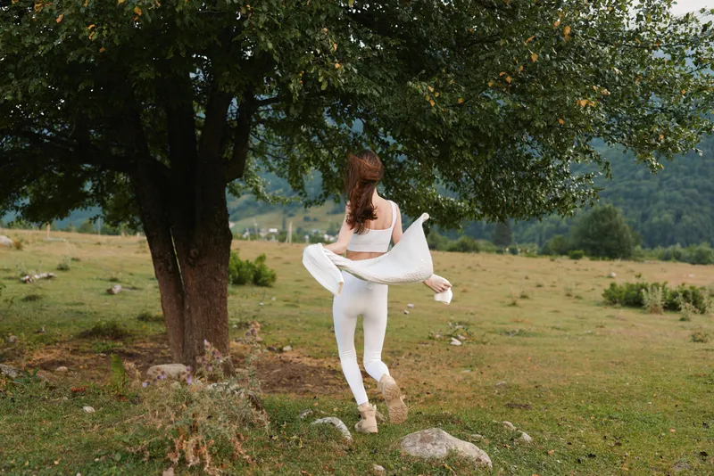 Young woman in a flowing white outfit spins joyfully in an open meadow beside a large tree, capturing a carefree outdoor moment
