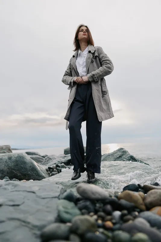 Young woman in a gray coat stands on rocky shore near calm sea under overcast sky. Fashion concept with casual outfit, natural environment, and moody atmosphere at the beach.