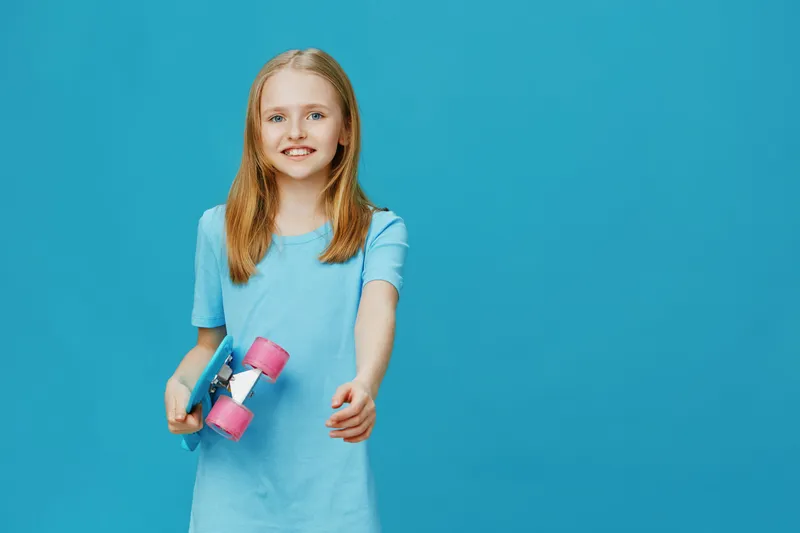 Cheerful young girl in a blue dress holding a skateboard, expressing joy against a bright turquoise background Perfect for seasonal sports and youth activities