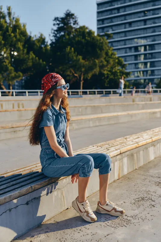 Lifestyle woman in modern boho-western denim street style sitting outdoors wearing sunglasses and bandana with film color tones, enjoying sunny urban environment.