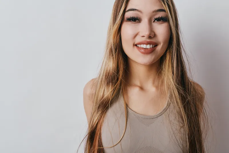Young woman with long hair smiling and looking at camera, wearing casual sleeveless top against plain light wall background for portrait and happiness.