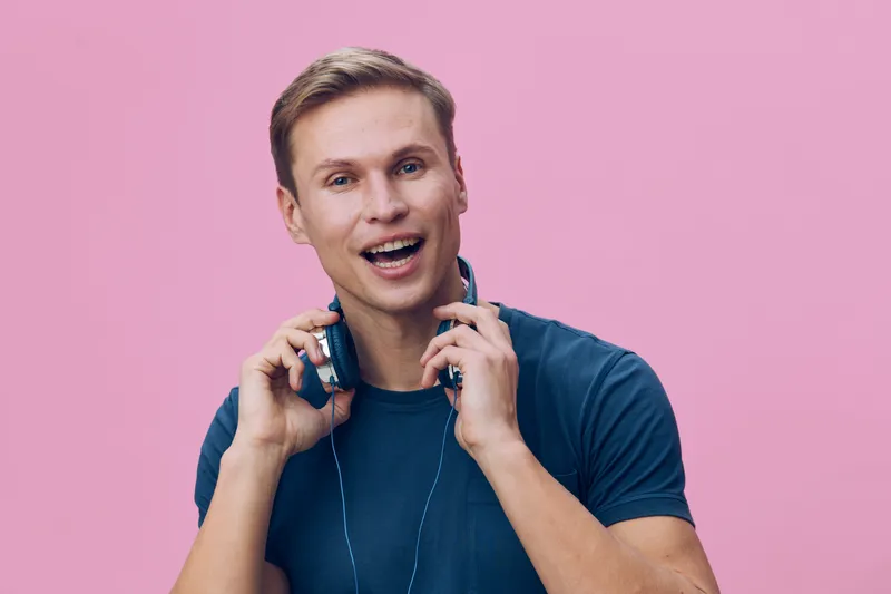 Upbeat young caucasian man wearing casual blue t-shirt and headphones, smiling happily while enjoying music, standing against pastel pink background. People leisure lifestyle concept, positive emotion