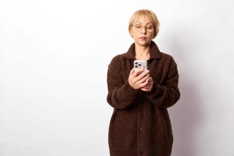 Senior woman holding smartphone for selfie in studio on white background, looking at screen with expression, portrait about modern technology and communication