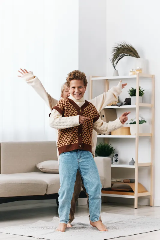 Joyful child dances in a bright living room, wearing a brown patterned vest, cream sweater, and blue jeans, capturing lively movement and carefree happiness.