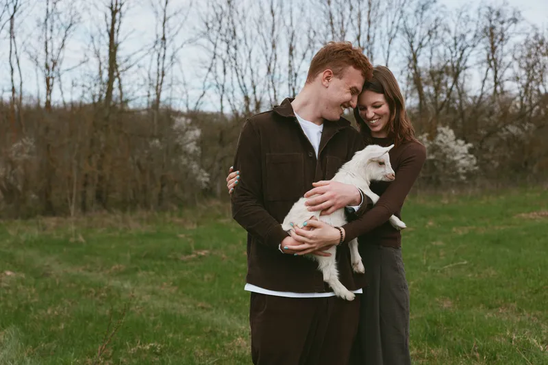 A joyful couple outdoors hug a small goat kid in a rural field, sharing a warm moment. A lifestyle scene highlighting affection, animal companionship, and countryside living.