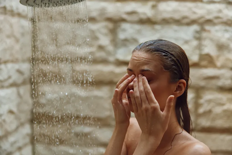 Woman washing face under outdoor shower with stone wall background, natural skincare routine, fresh water droplets in warm sunlight, serene wellness moment.