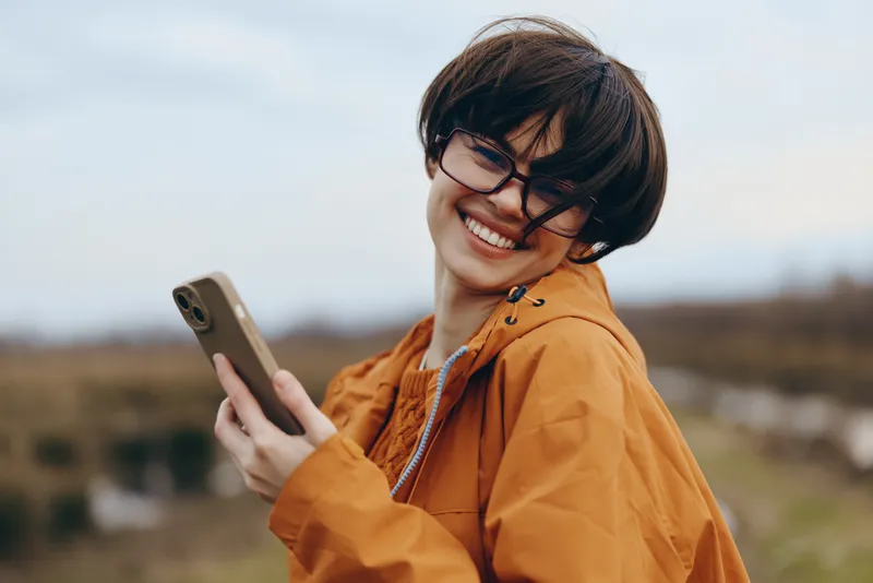 Smiling woman lifestyle outdoors, orange jacket, holding smartphone near her face, laughing while reading messages, blurred countryside landscape behind, natural daylight, casual travel portrait,