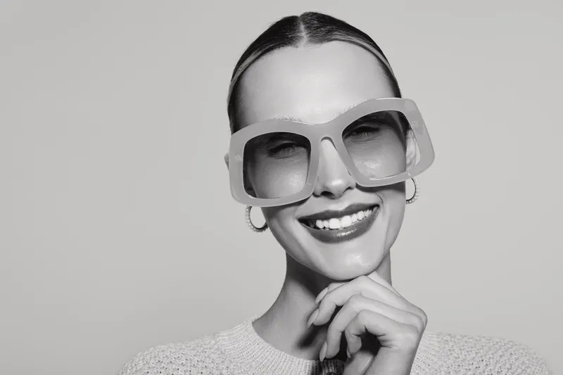 Smiling light skinned Caucasian woman with slicked hair wearing oversized sunglasses and white sweater in studio.