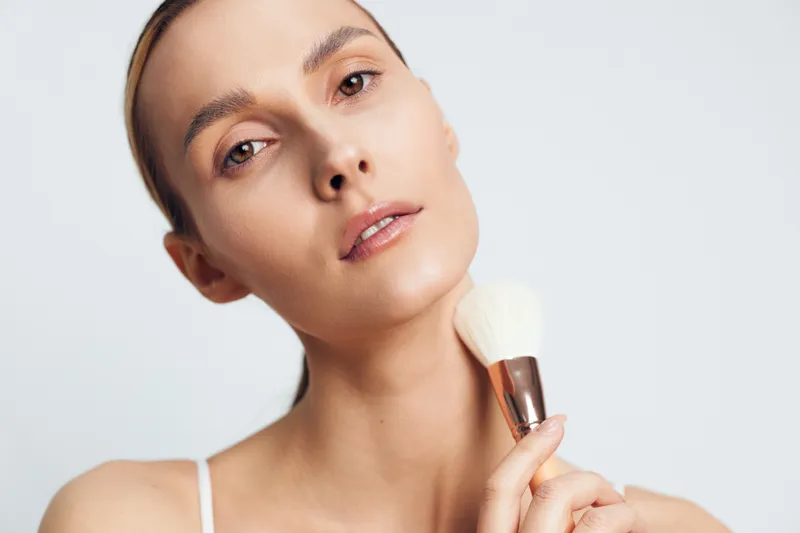 Young woman with clear skin gently applies makeup with a brush while looking at the camera against a plain white background in a minimalistic beauty portrait.