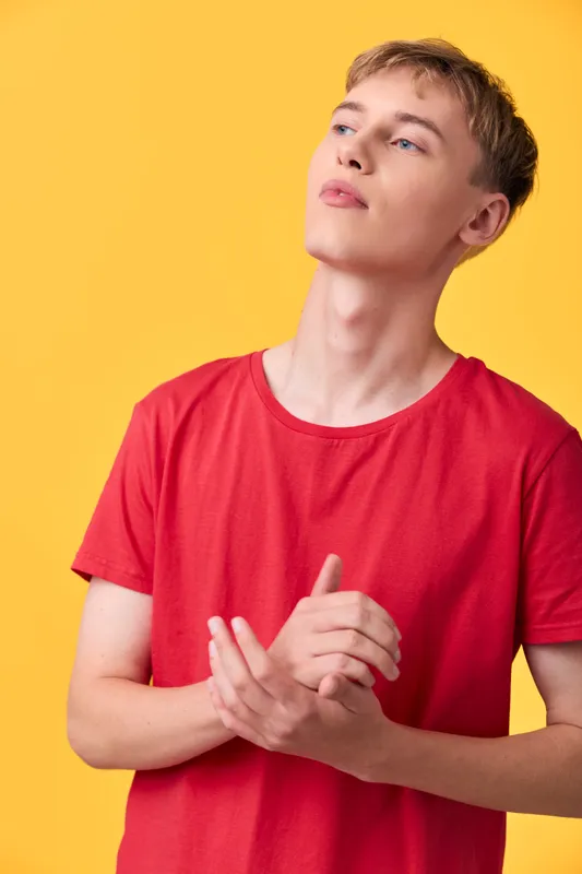 Young man in a red shirt stands against a bright yellow background, a contemplative pose with clasped hands, casual fashion vibe, studio shot, clean composition, modern mood, bold color contrast,