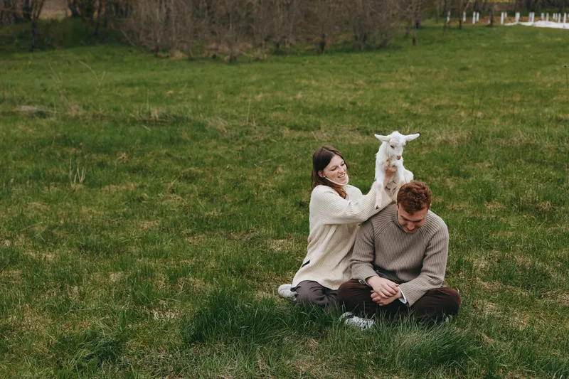 A smiling couple sits on green grass in a field with a playful goat kid nearby, enjoying a relaxed moment together. Natural light and candid lifestyle vibe convey warmth, companionship, and simple