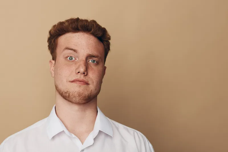 Portrait of a young man with red hair and freckles wearing a white shirt against beige background. Concept of natural beauty, confidence, and casual style for lifestyle and fashion.